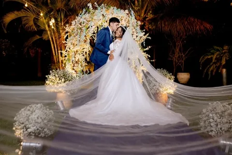 Casal de noivos sob arco de flores iluminado à noite, véu longo estendido